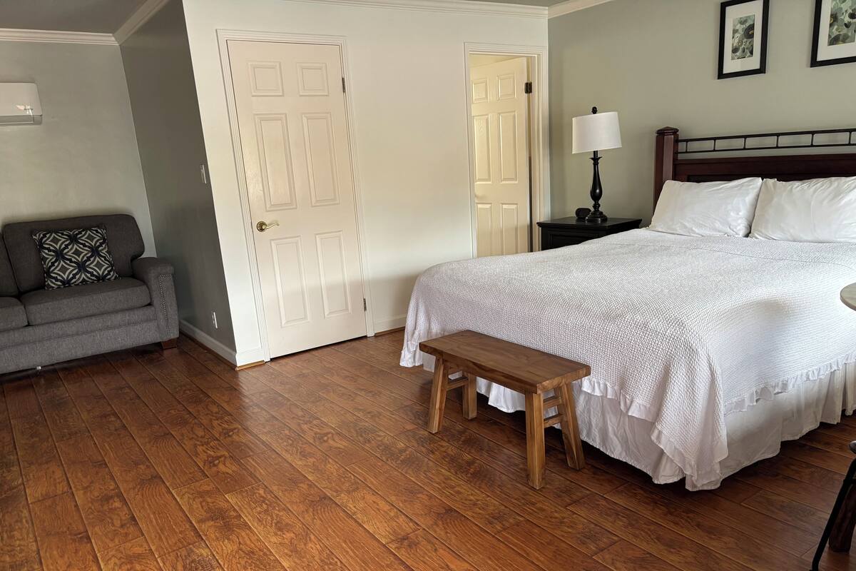 A bedroom with a large bed, a bench, a gray sofa, and two doors.