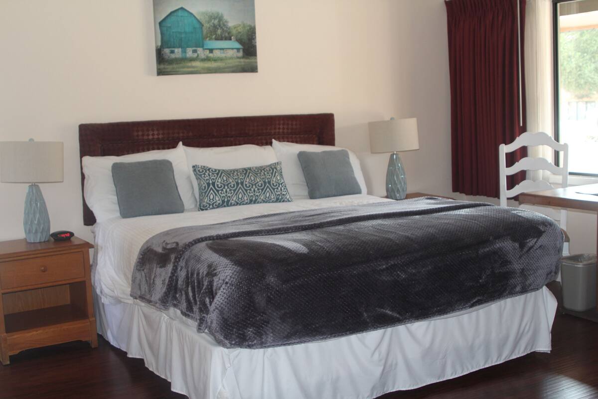 A neatly made bed with a dark blanket, two bedside tables with lamps, a chair, and a painting of a barn on the wall.