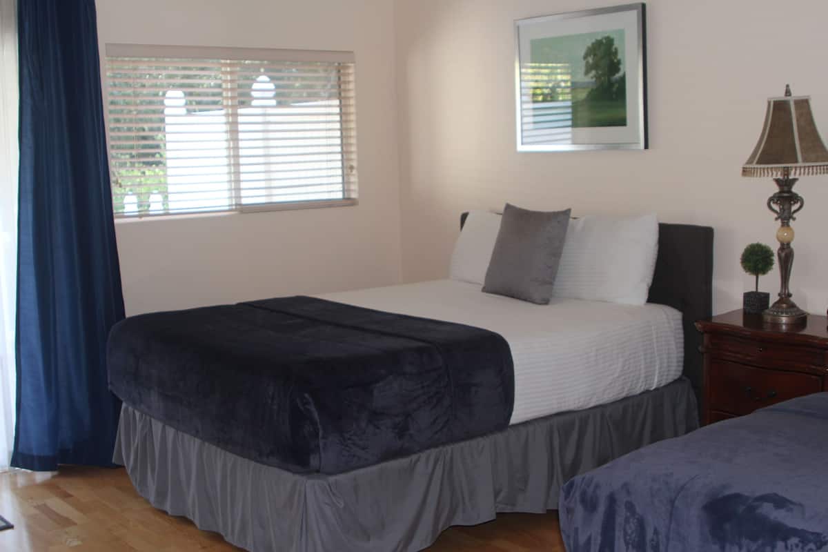 A bedroom with a bed, a nightstand, a lamp, a window with blinds, and a framed picture on the wall.