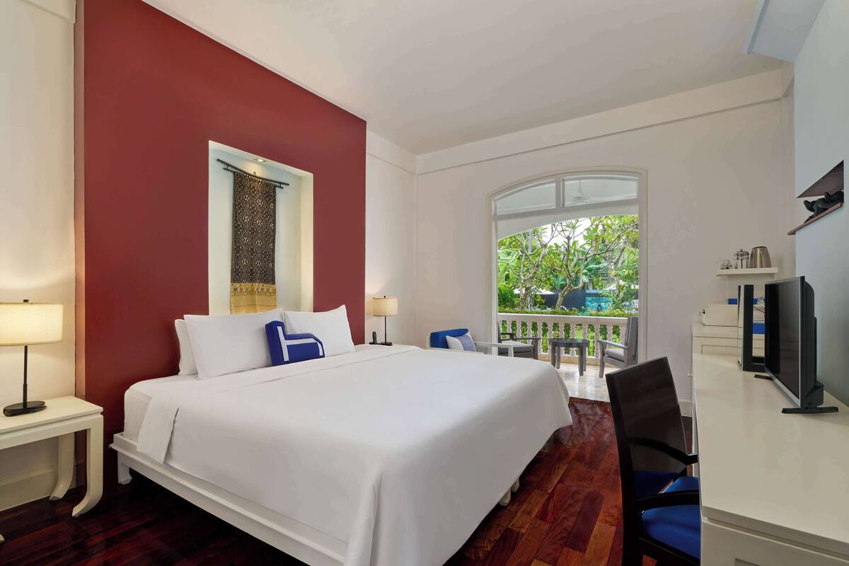 Une chambre d’hôtel avec un grand lit, un bureau avec une télévision, une fenêtre donnant sur un espace verdoyant et un balcon.