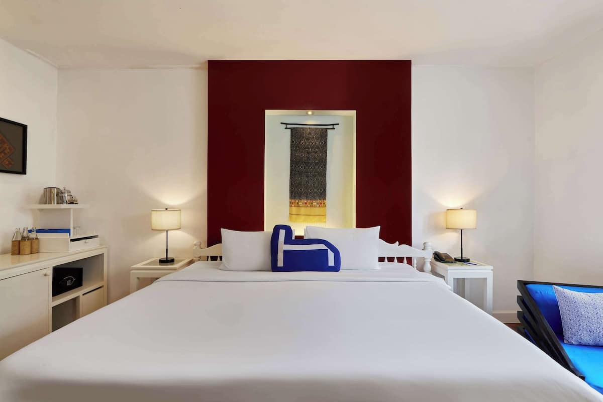 Une chambre d’hôtel avec un grand lit, deux tables de chevet avec des lampes, un petit bureau avec une lampe et un canapé bleu.