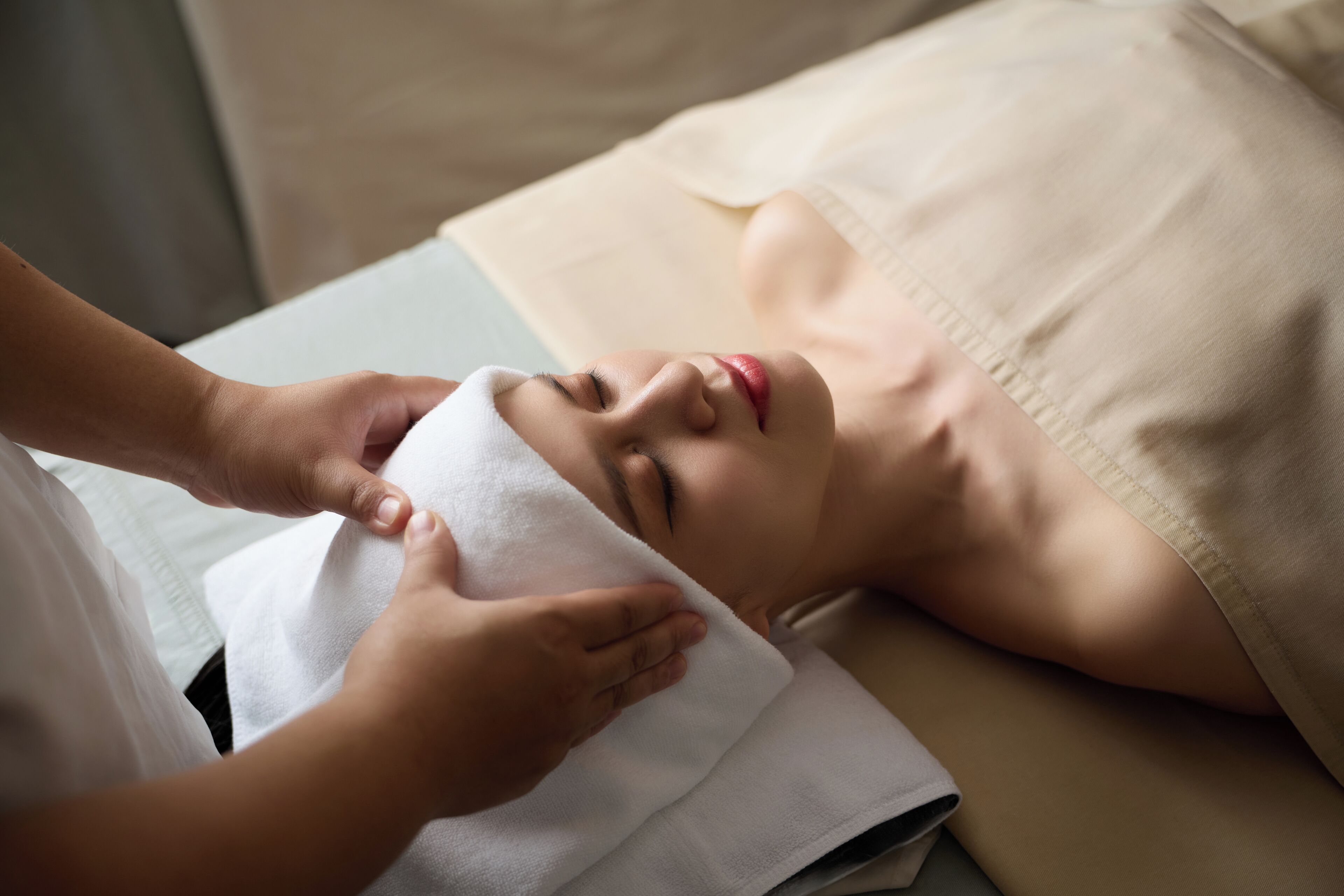 A person lying on a bed with a white towel over their head, receiving a facial treatment.