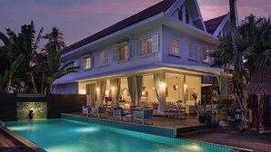 Un edificio de dos pisos con piscina, mobiliario exterior y vegetación tropical.