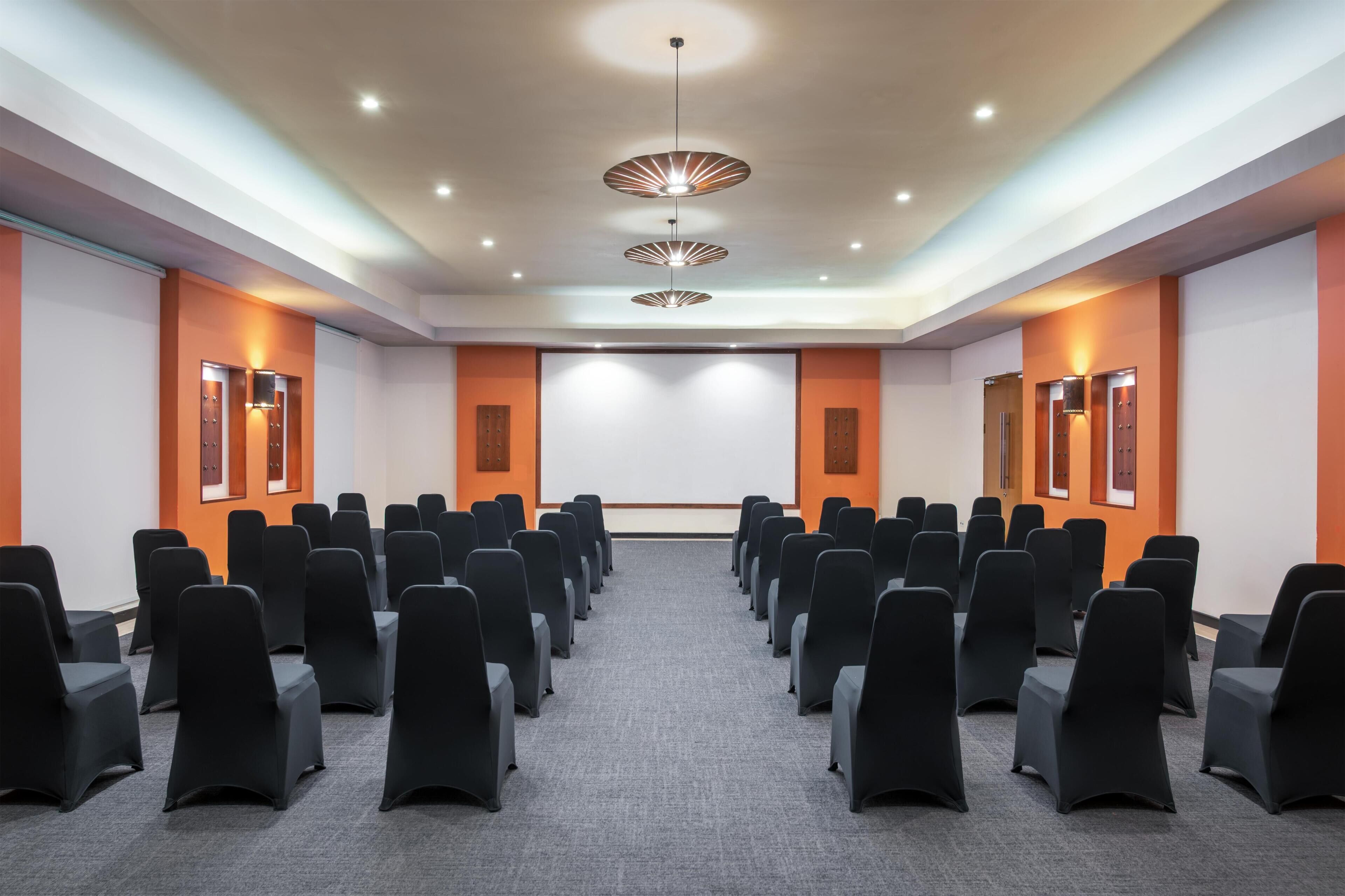 A conference room with rows of black chairs, a projector screen, and three pendant lights.