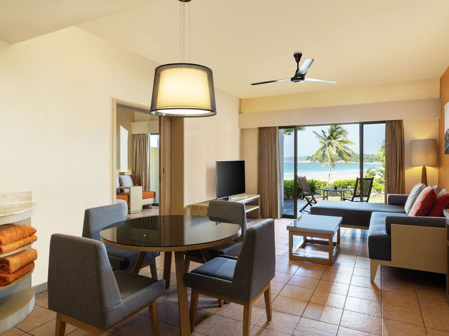 A hotel room with a dining area, a sofa, a TV, and a balcony with a view of the beach.