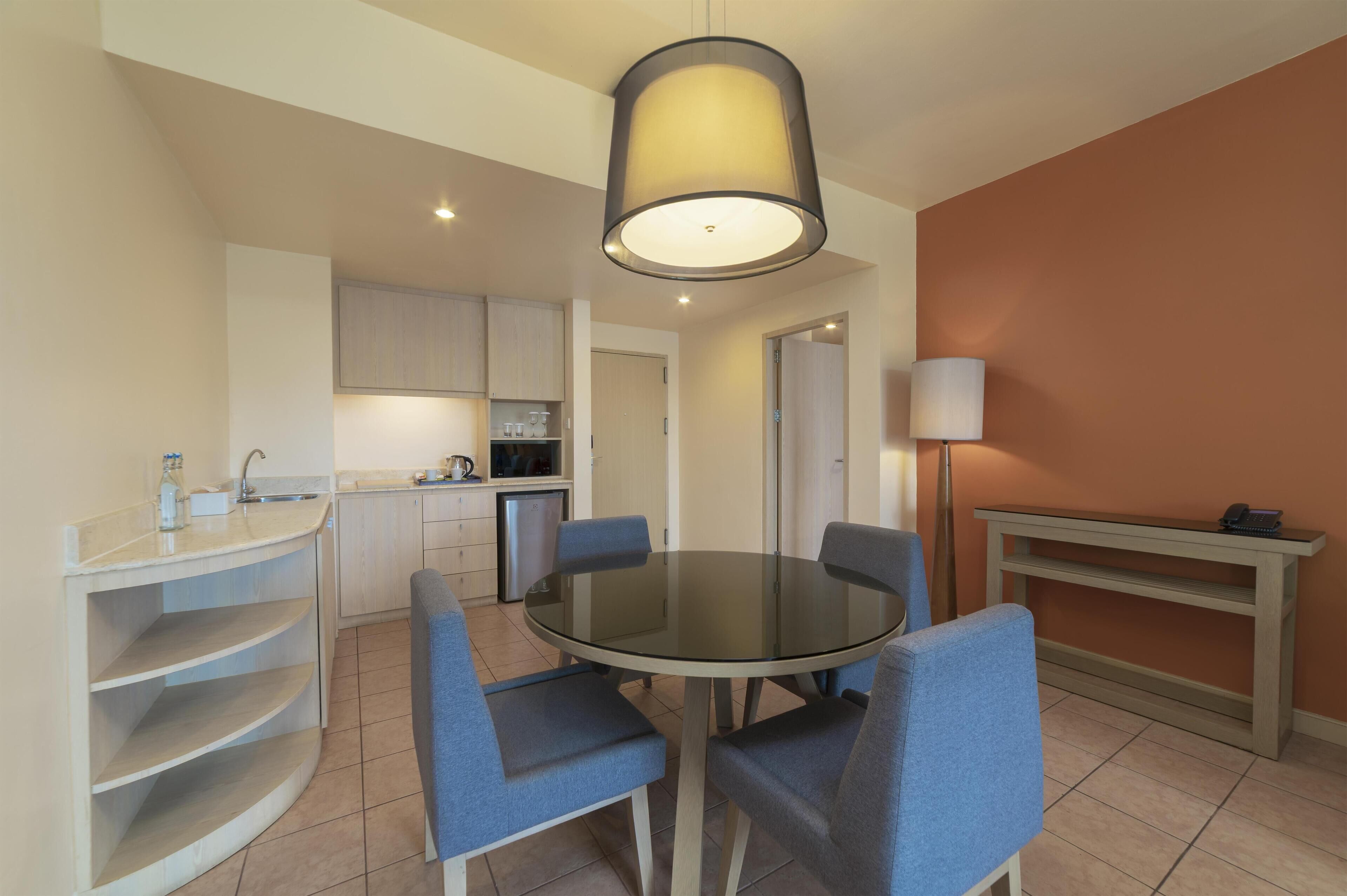 A modern kitchen with a round glass table and blue chairs, a built-in shelf, and a pendant light.