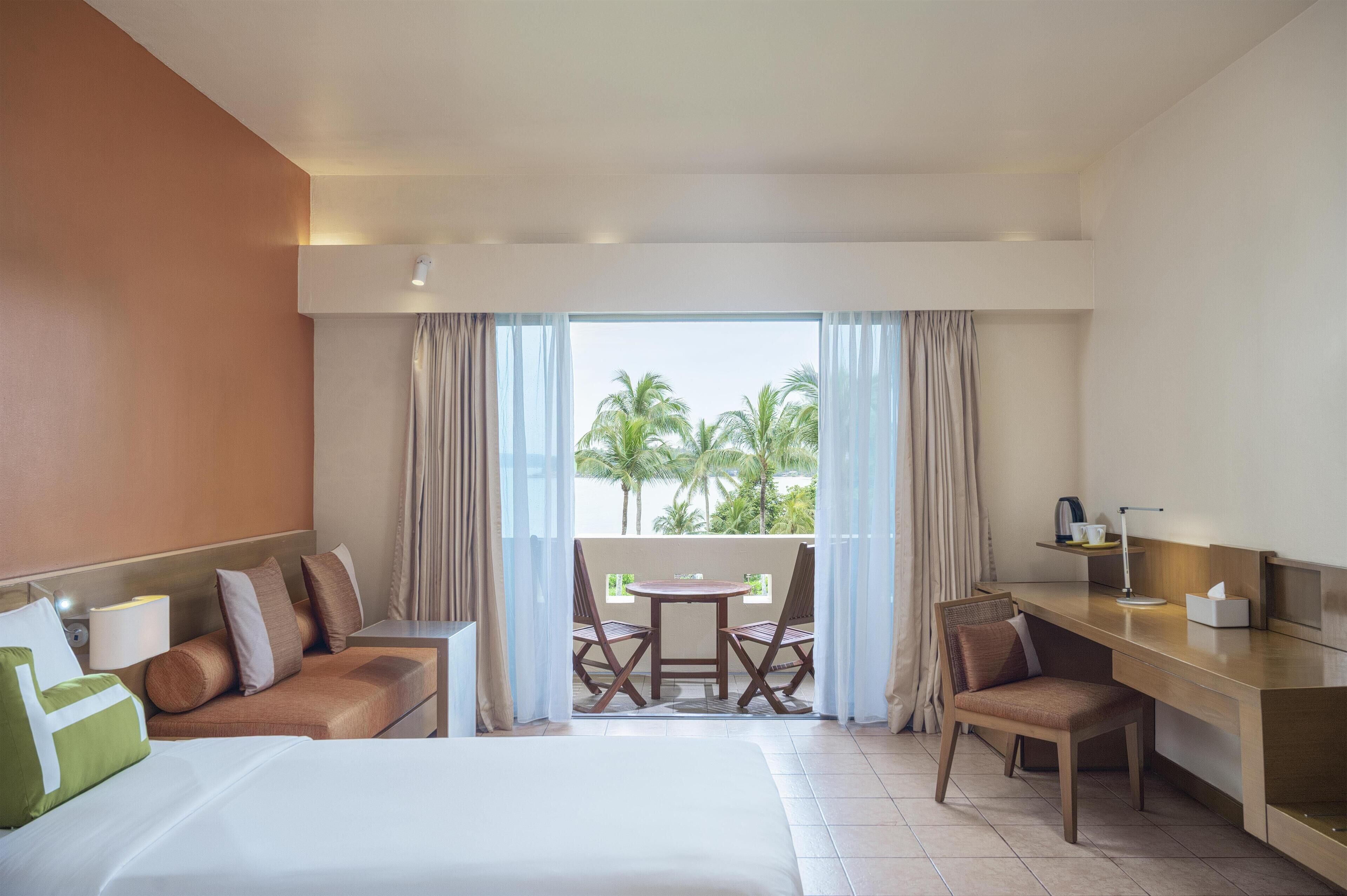 Ein Hotelzimmer mit Bett, Schreibtisch, Stuhl, kleinem Tisch und einem Balkon mit Blick auf Palmen.
