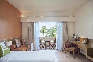 A hotel room with a bed, a desk, a chair, a small table, and a balcony with a view of palm trees. - Homm Laguna Bintan (Bintan)