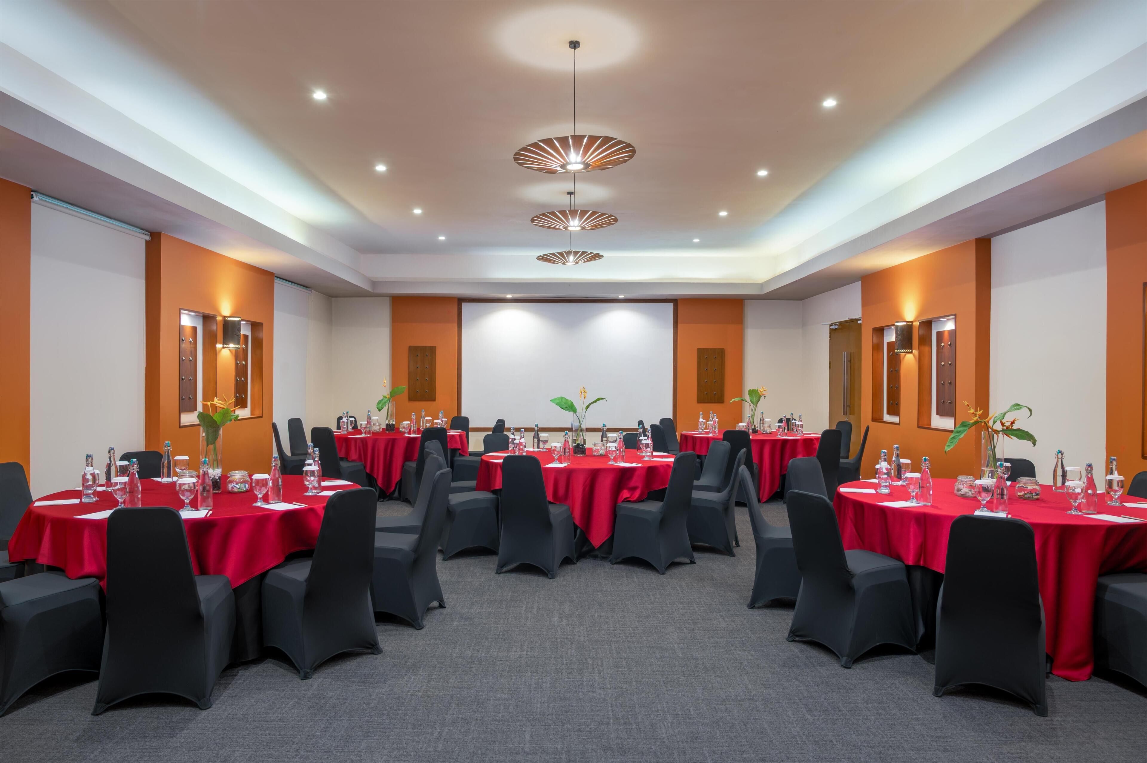 A conference room with multiple tables, black chairs, red tablecloths, and a projector screen.