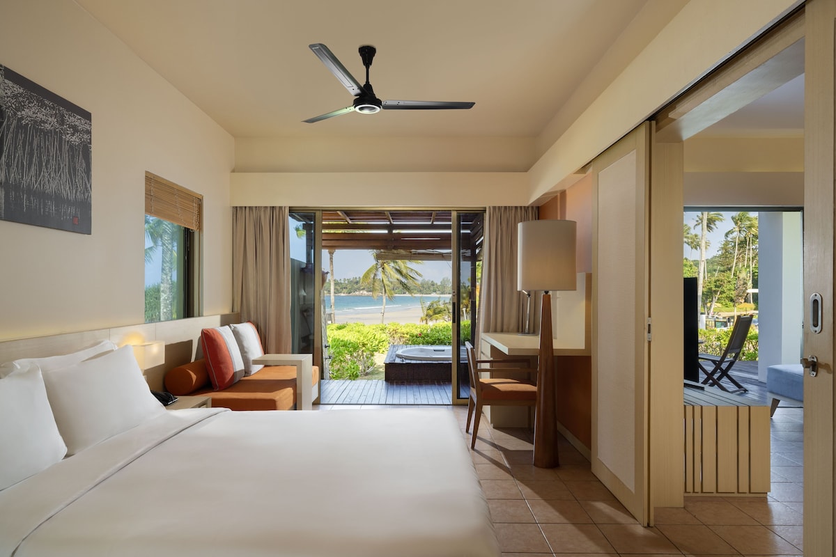 Quarto de hotel com cama, escrivaninha, cadeira, ventoinha no teto e vista para a praia.