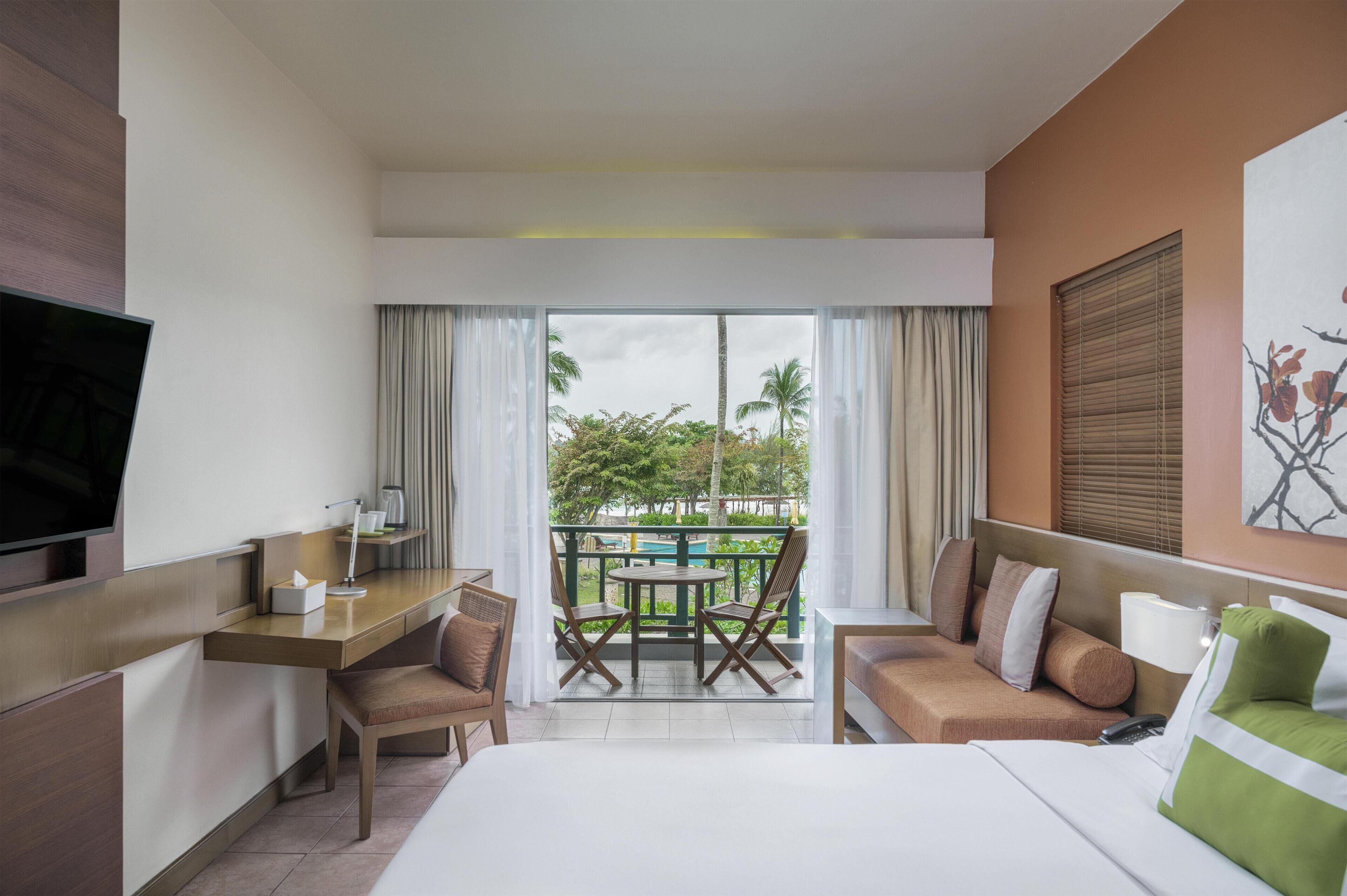 Habitación de hotel con cama, escritorio, silla, sofá, televisor y balcón con vista a palmeras y una piscina.