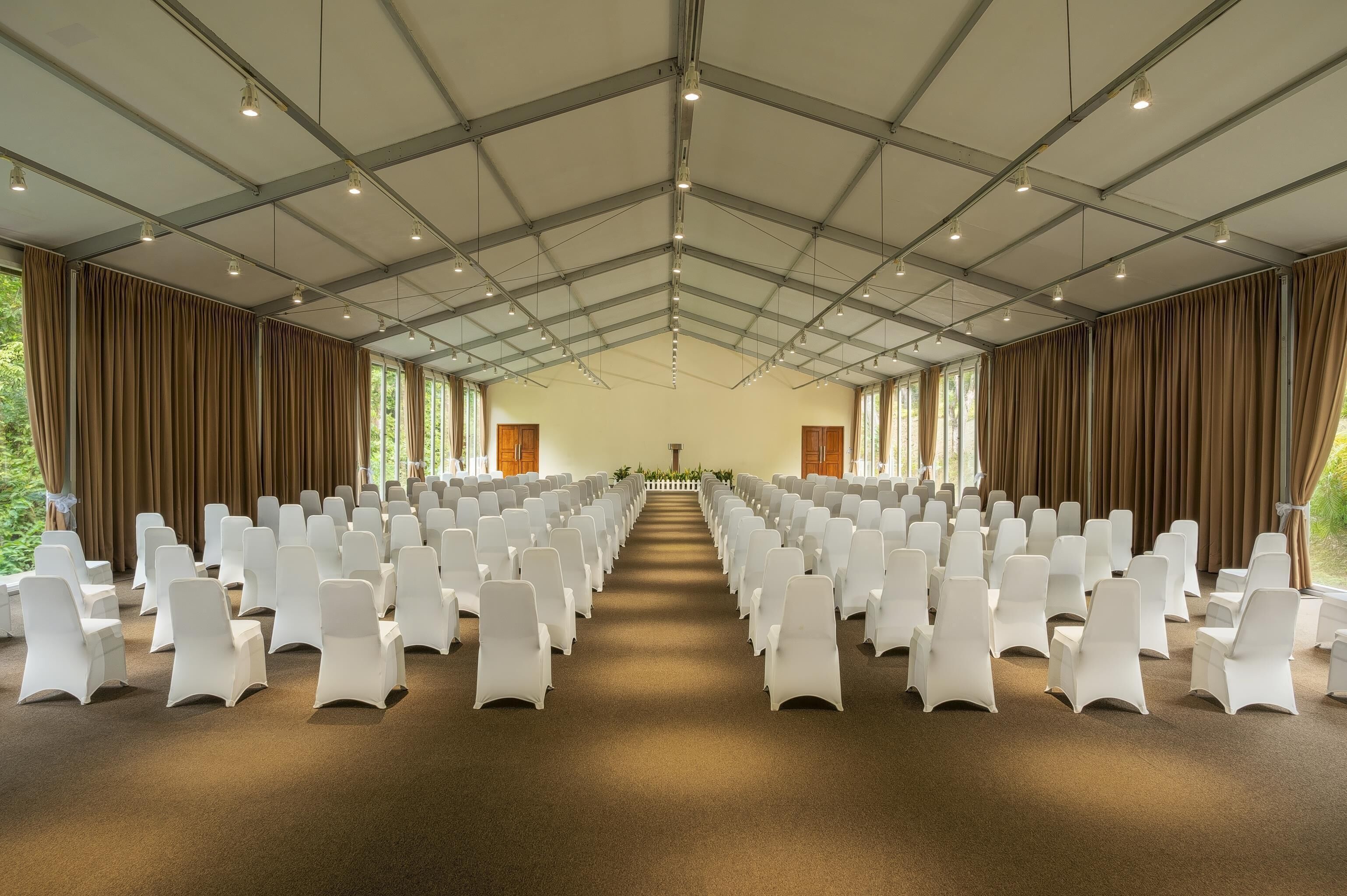 A large hall with rows of white chairs and brown curtains, a high ceiling with lights, and large windows.
