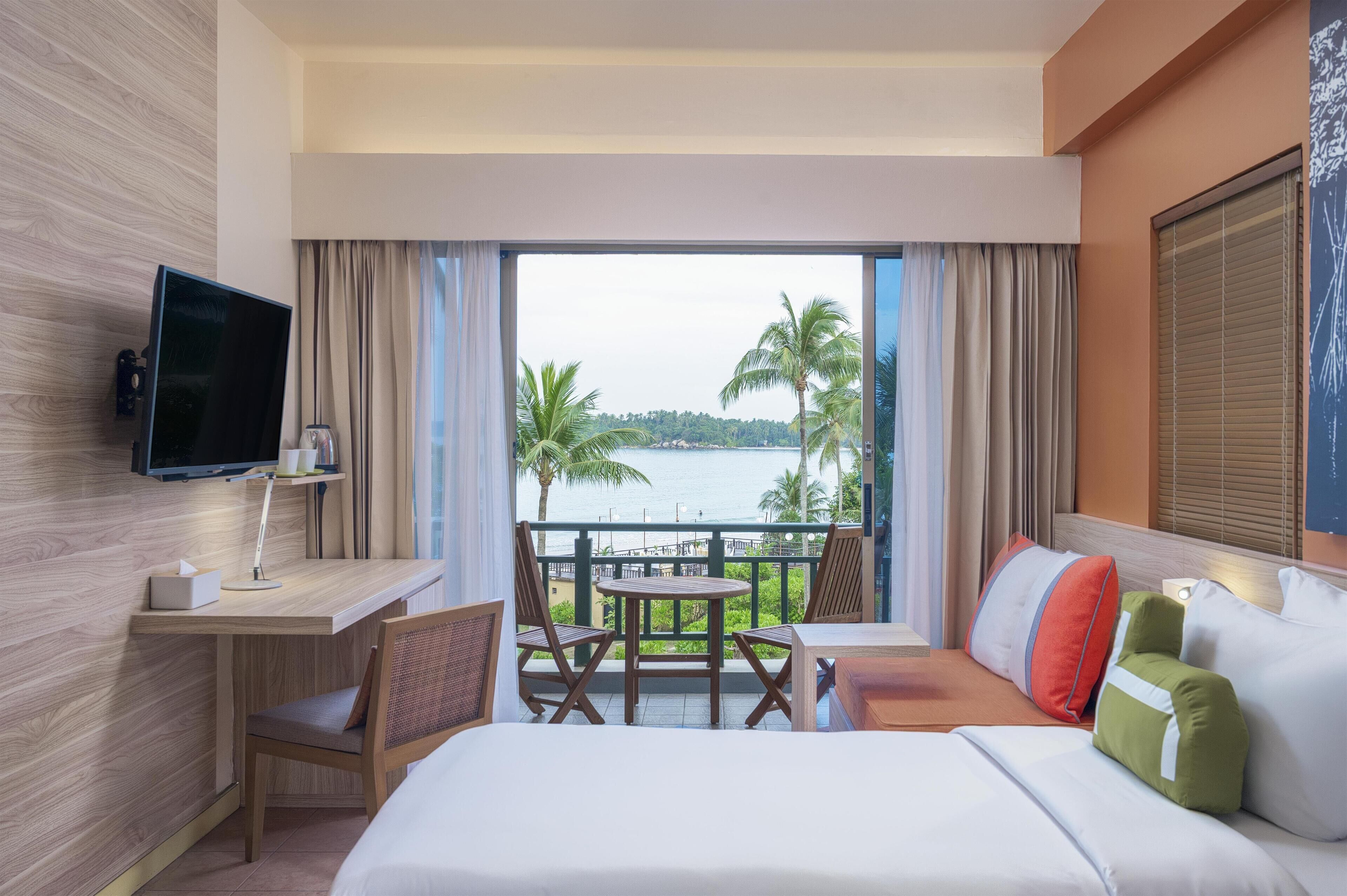 A hotel room with a bed, a desk, a TV, a table with chairs, and a balcony with a view of the sea and palm trees.