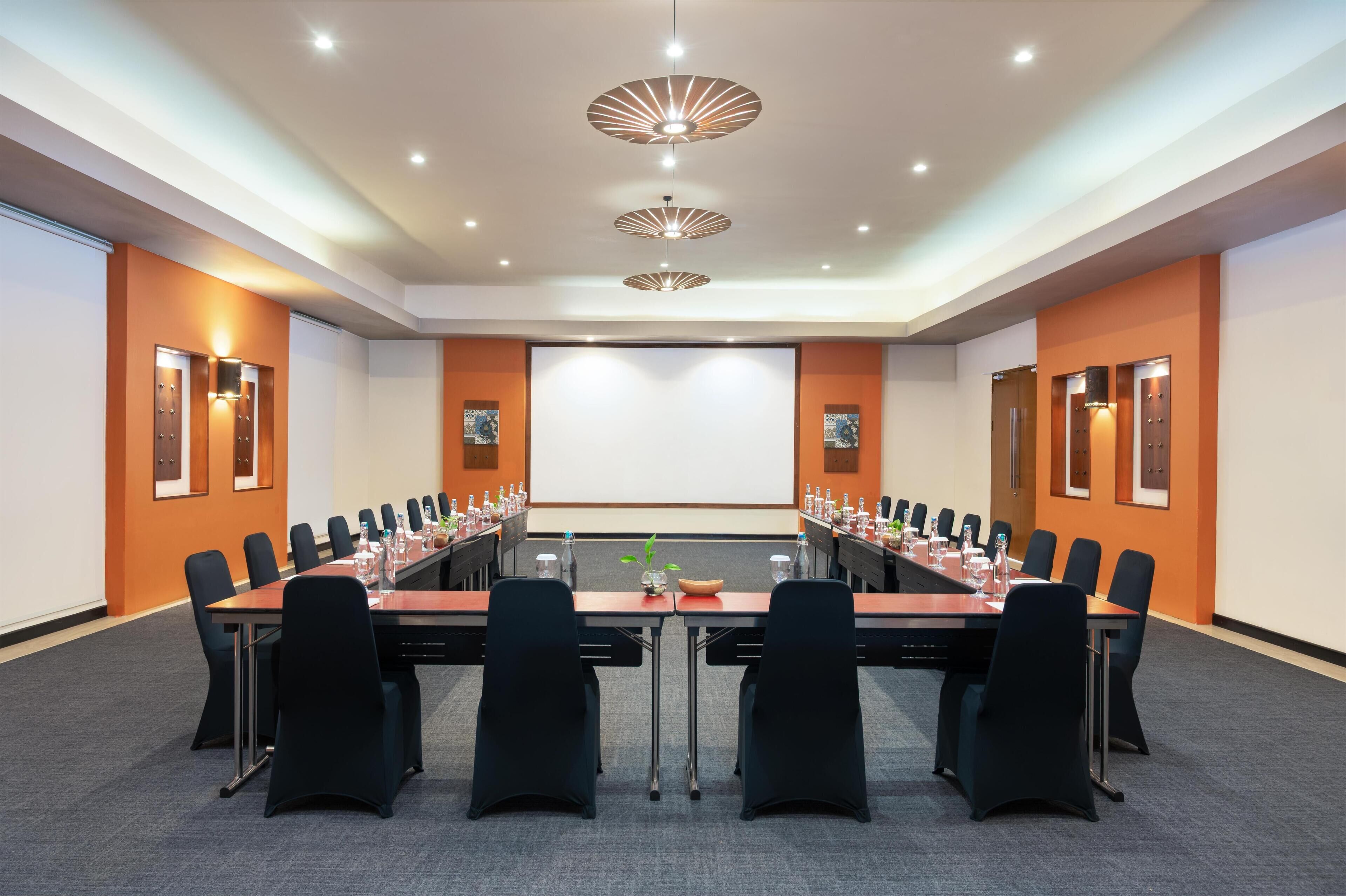 Een vergaderzaal met een lange tafel, zwarte stoelen en een projectiescherm.