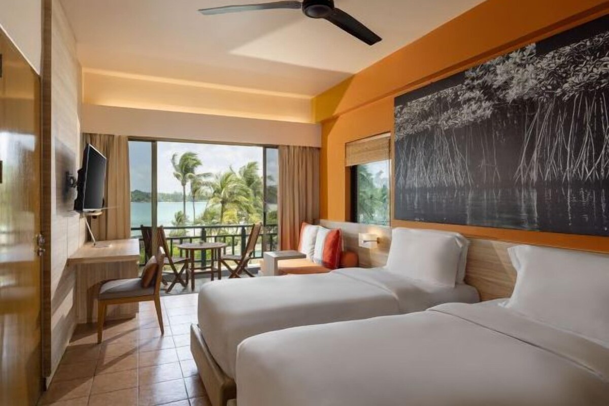 Habitación de hotel con dos camas, un escritorio, una silla, televisión y balcón con vistas a palmeras.