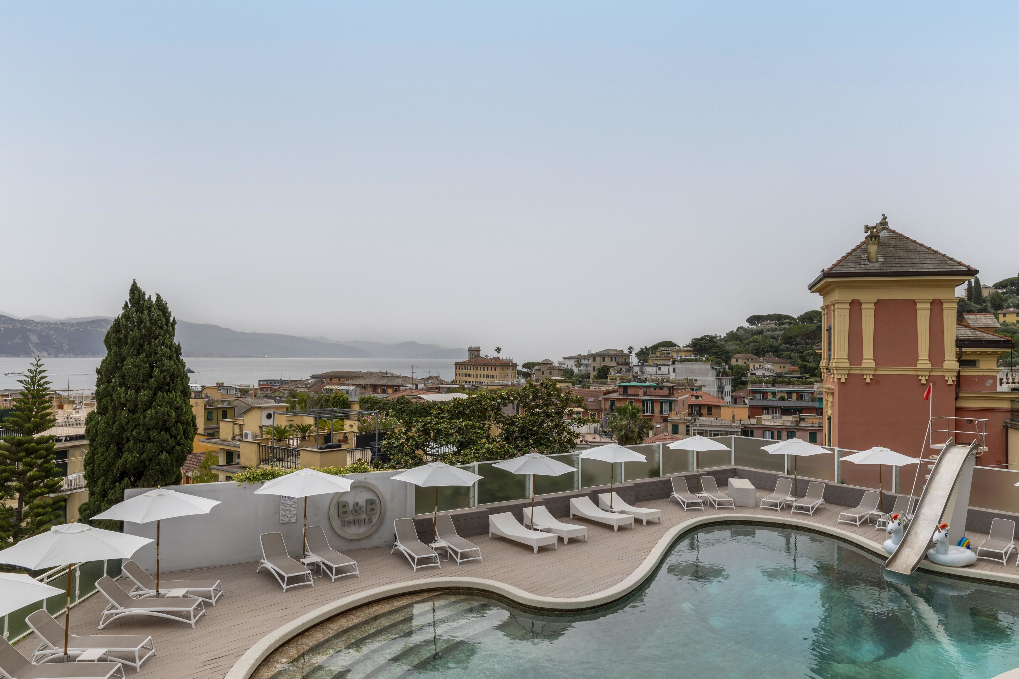 A rooftop pool area with lounge chairs and umbrellas, overlooking a cityscape and the sea.