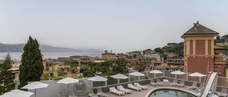A rooftop pool area with lounge chairs and umbrellas, overlooking a cityscape and the sea.