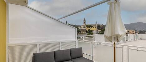 A balcony with a grey sofa, a white umbrella, and a view of buildings and mountains.