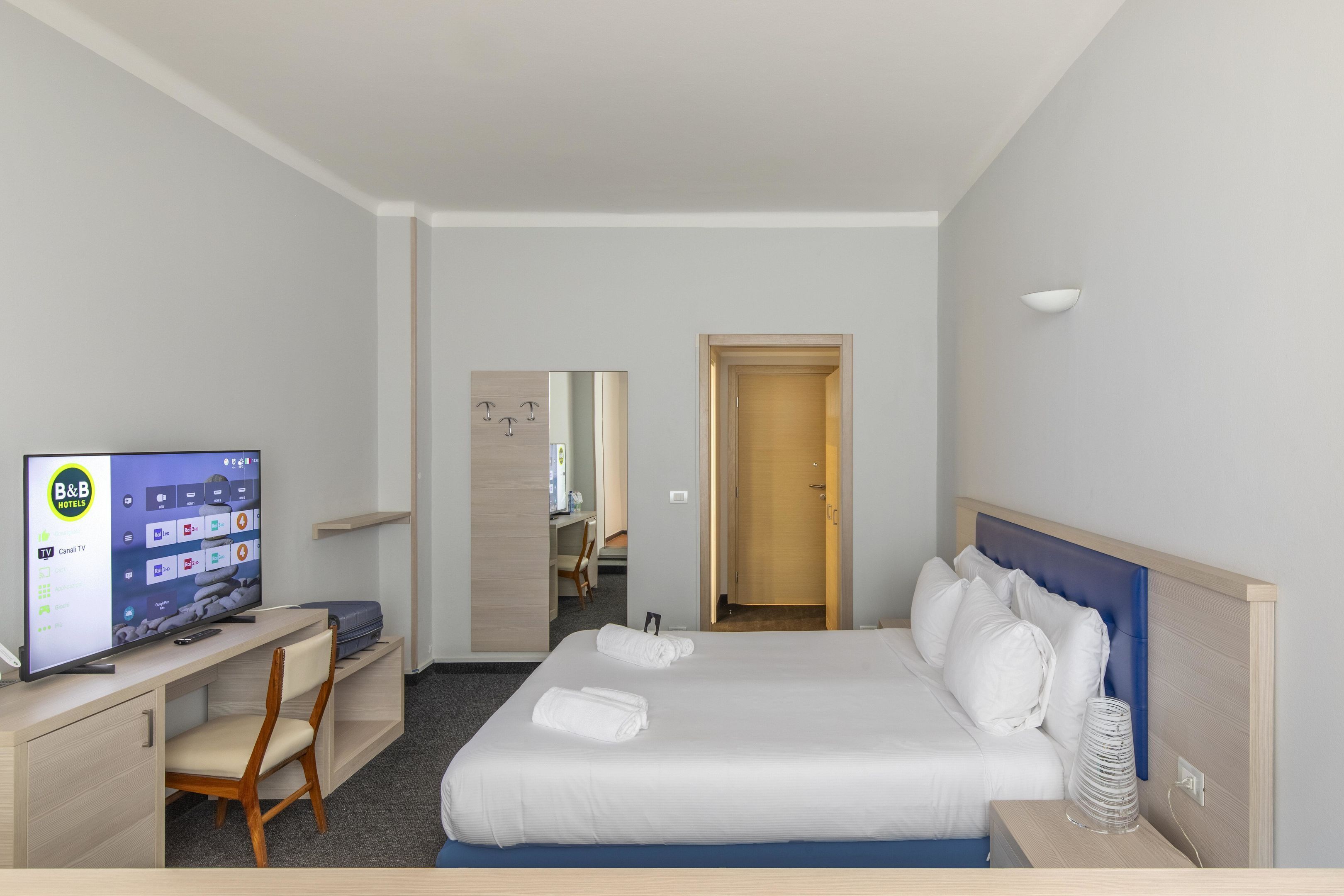 A hotel room with a bed, a desk with a TV, a chair, and a door leading to a bathroom.
