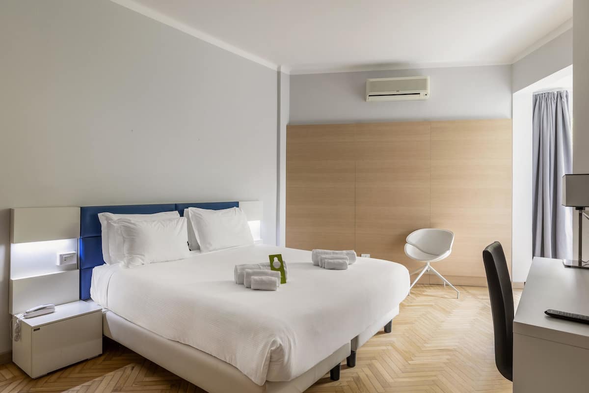 A hotel room with a large bed, a desk, a chair, and a small bedside table.
