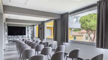 A conference room with rows of chairs, a large flat-screen TV, and a view of buildings and trees through large windows.