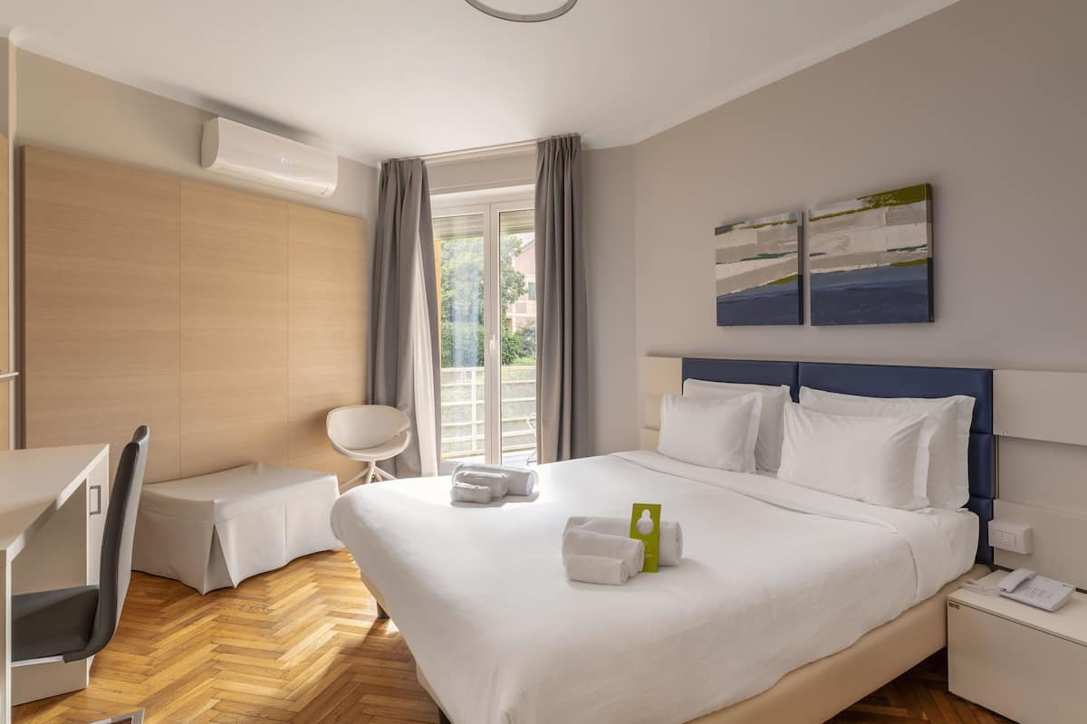 A hotel room with a bed, a desk, a chair, a small table, a nightstand, and a window with curtains.