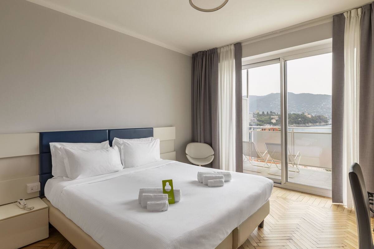 A hotel room with a large bed, a view of the city through a balcony, and a chair outside.