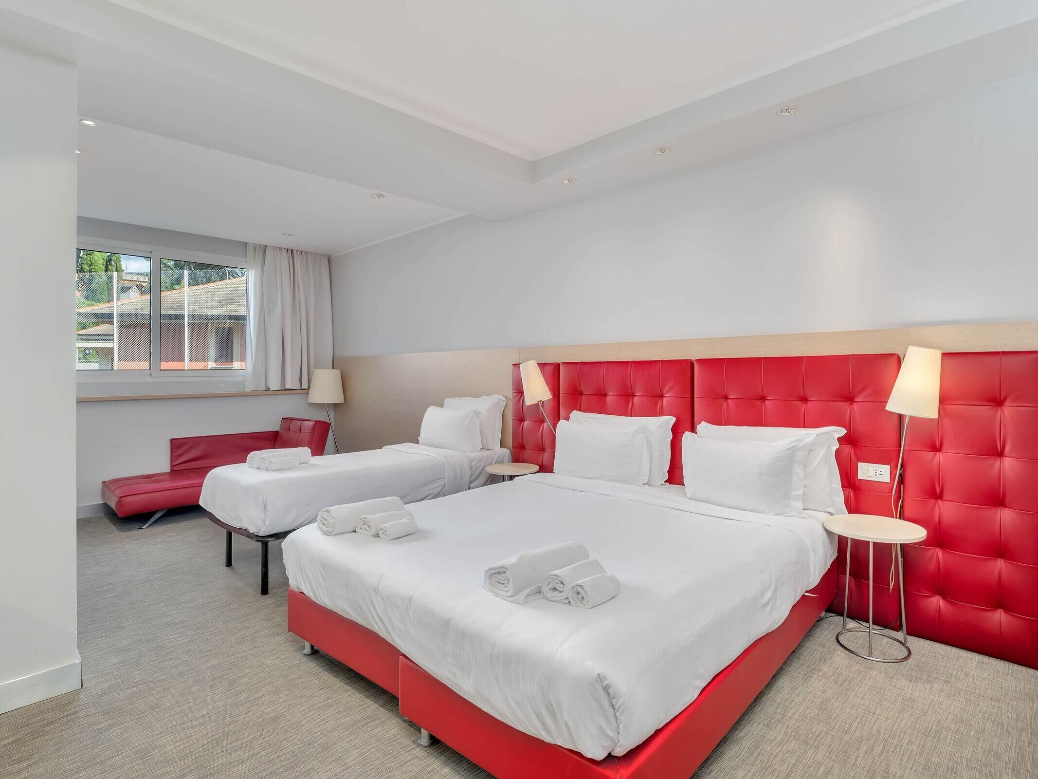 Habitación de hotel con dos camas, cabecera y silla rojas, una mesita y una ventana con cortinas.