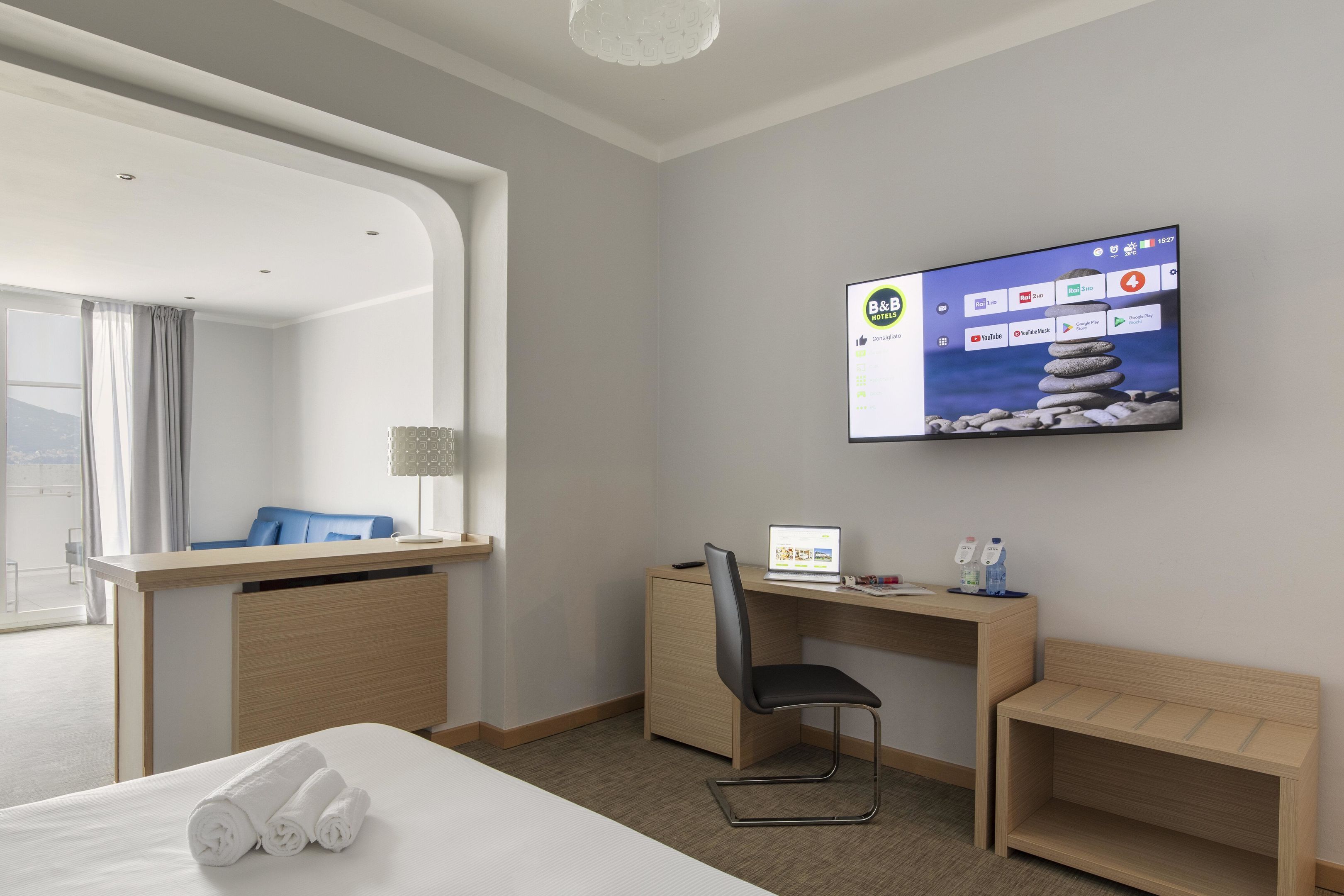 Habitación de hotel con una cama, un escritorio con una computadora portátil, una silla, un televisor en la pared y vistas al exterior.