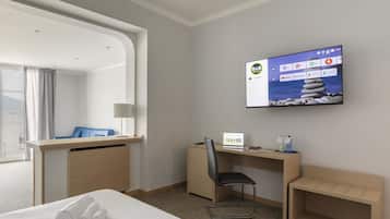 Quarto de hotel com cama, escrivaninha com laptop, cadeira, TV na parede e vista para o exterior.