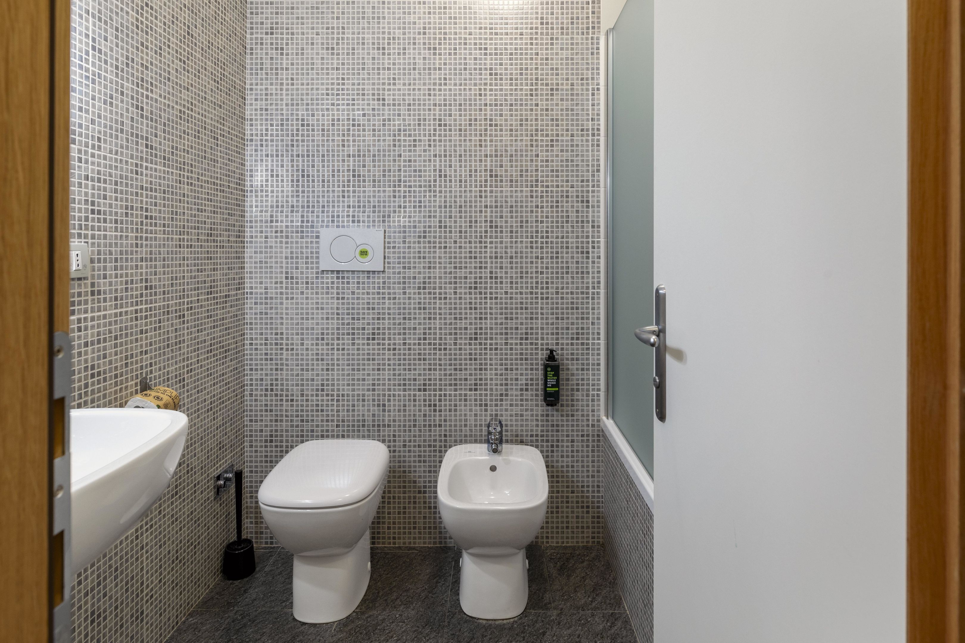 A bathroom with a white sink, toilet, and bidet, tiled walls, and a door with a handle.
