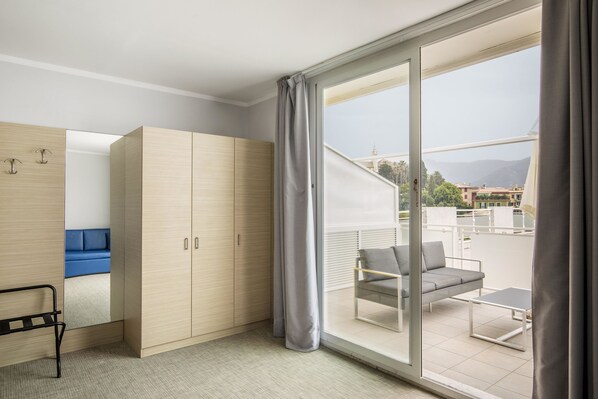 Una habitación de hotel moderna con un gran ventanal corredero al balcón, un sofá, una silla y una cama con cabecera azul.