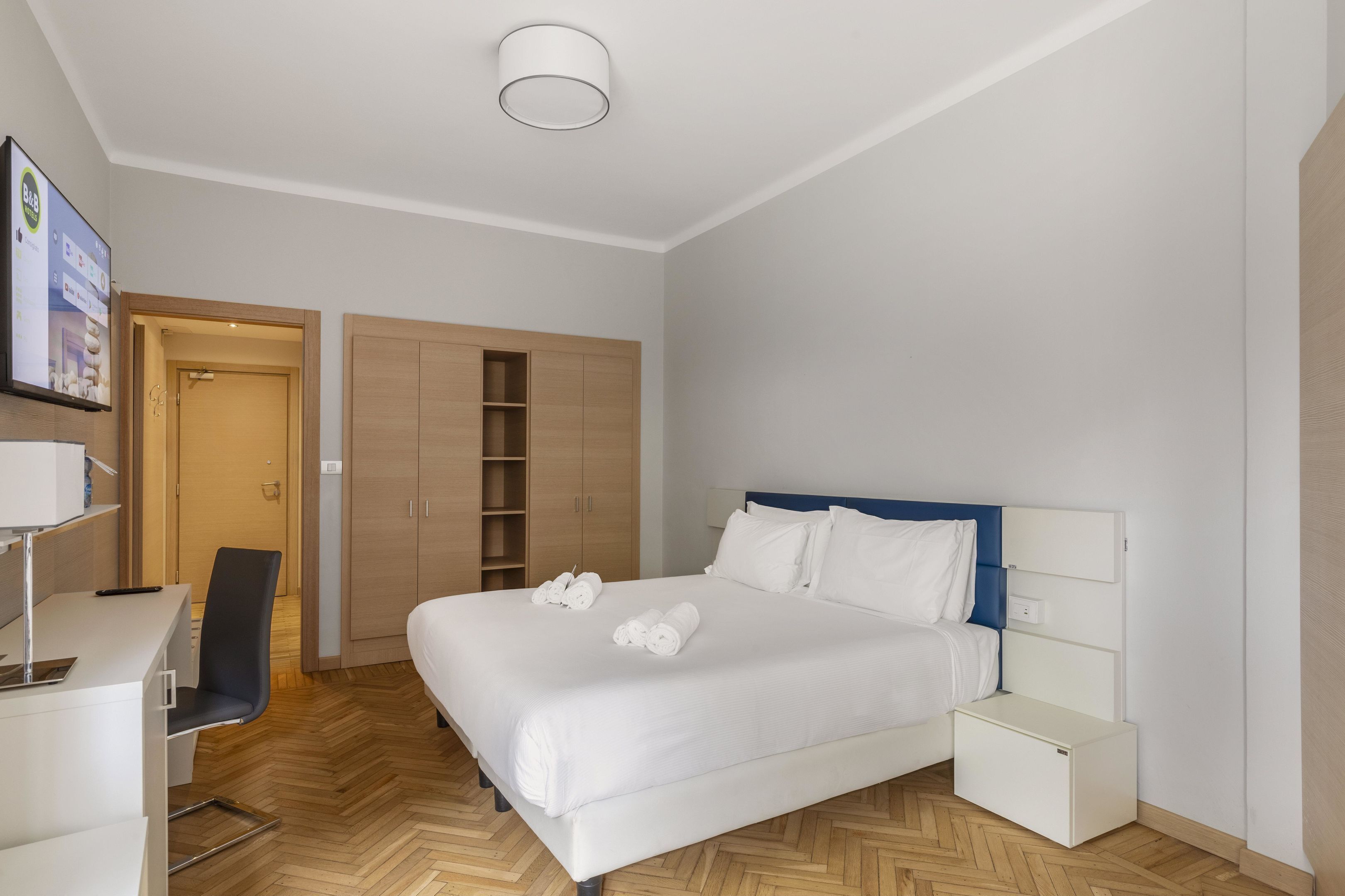 A modern hotel room with a large bed, a desk, a chair, and a wardrobe.