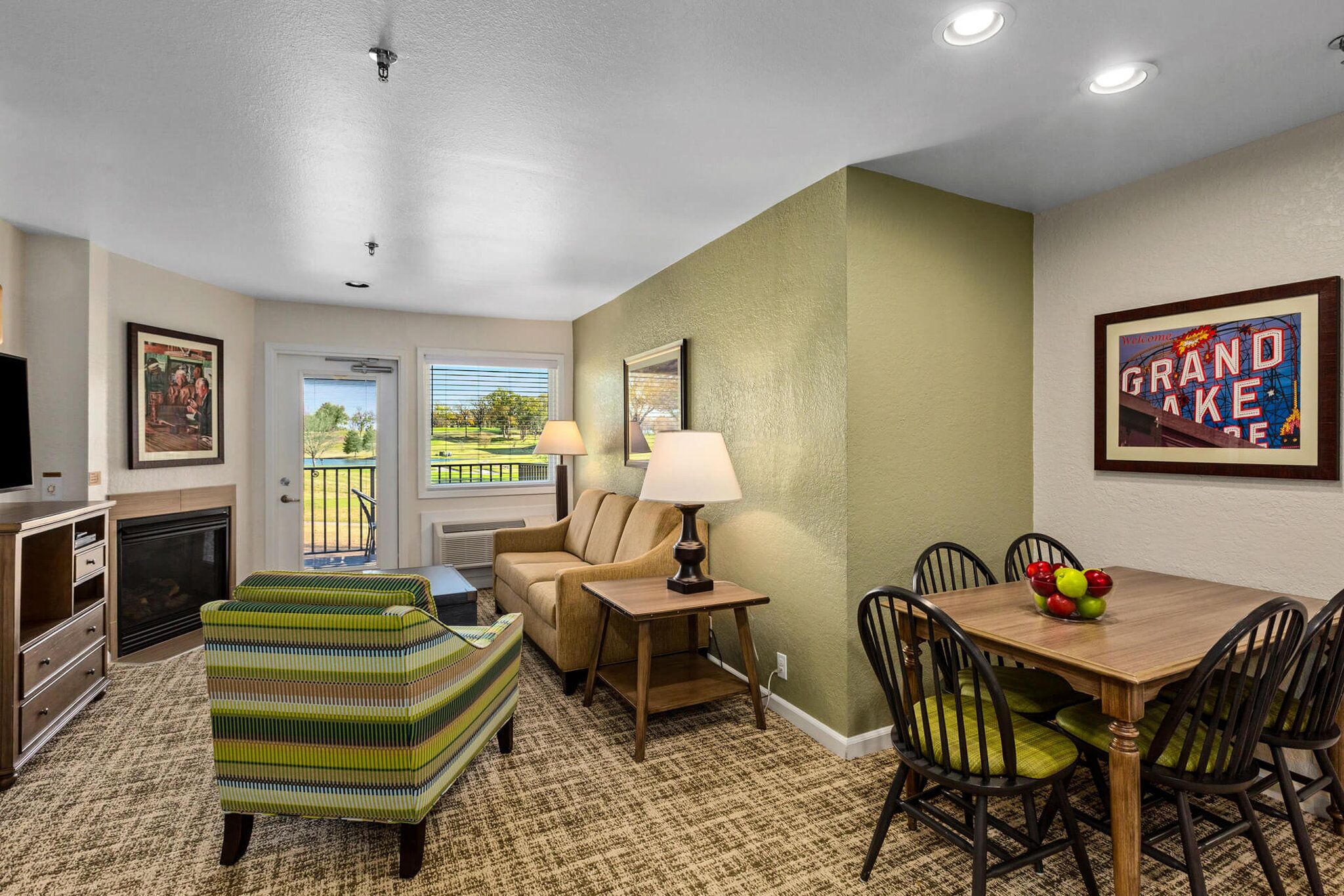 A cozy living room with a sofa, armchair, and a dining table with chairs. There is a television, a fireplace, and a window with blinds.