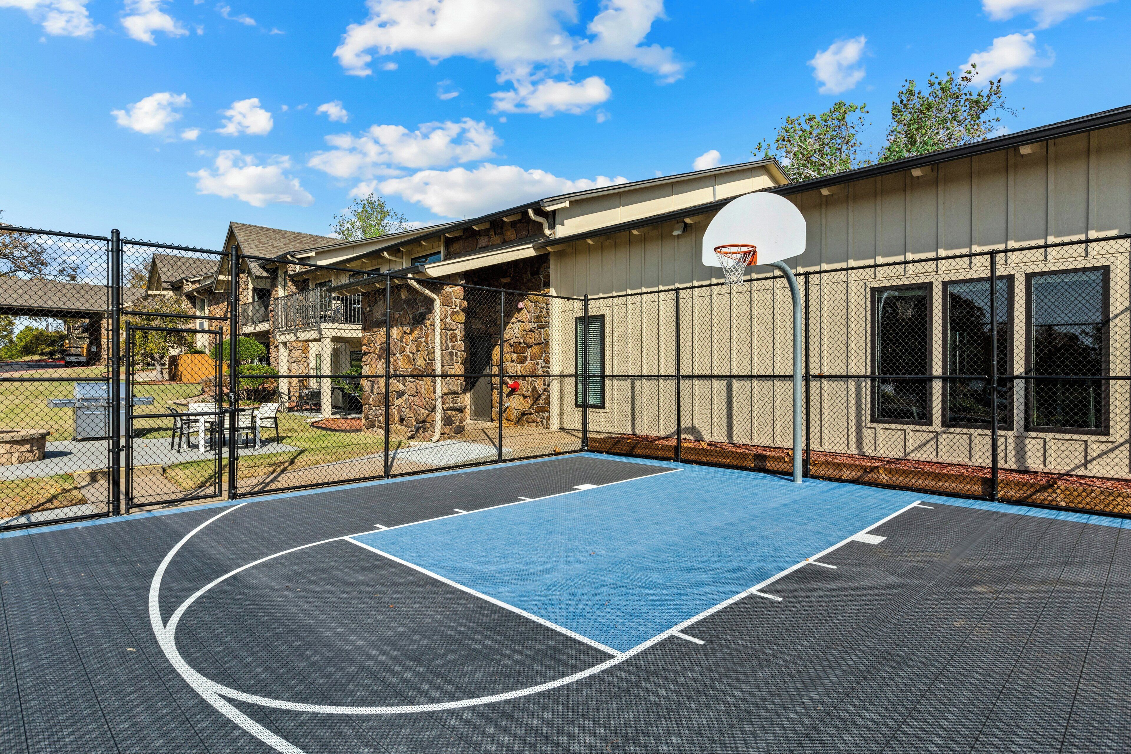 An outdoor basketball court with a chain-link fence, a basketball hoop, and a building with a stone facade in the background.