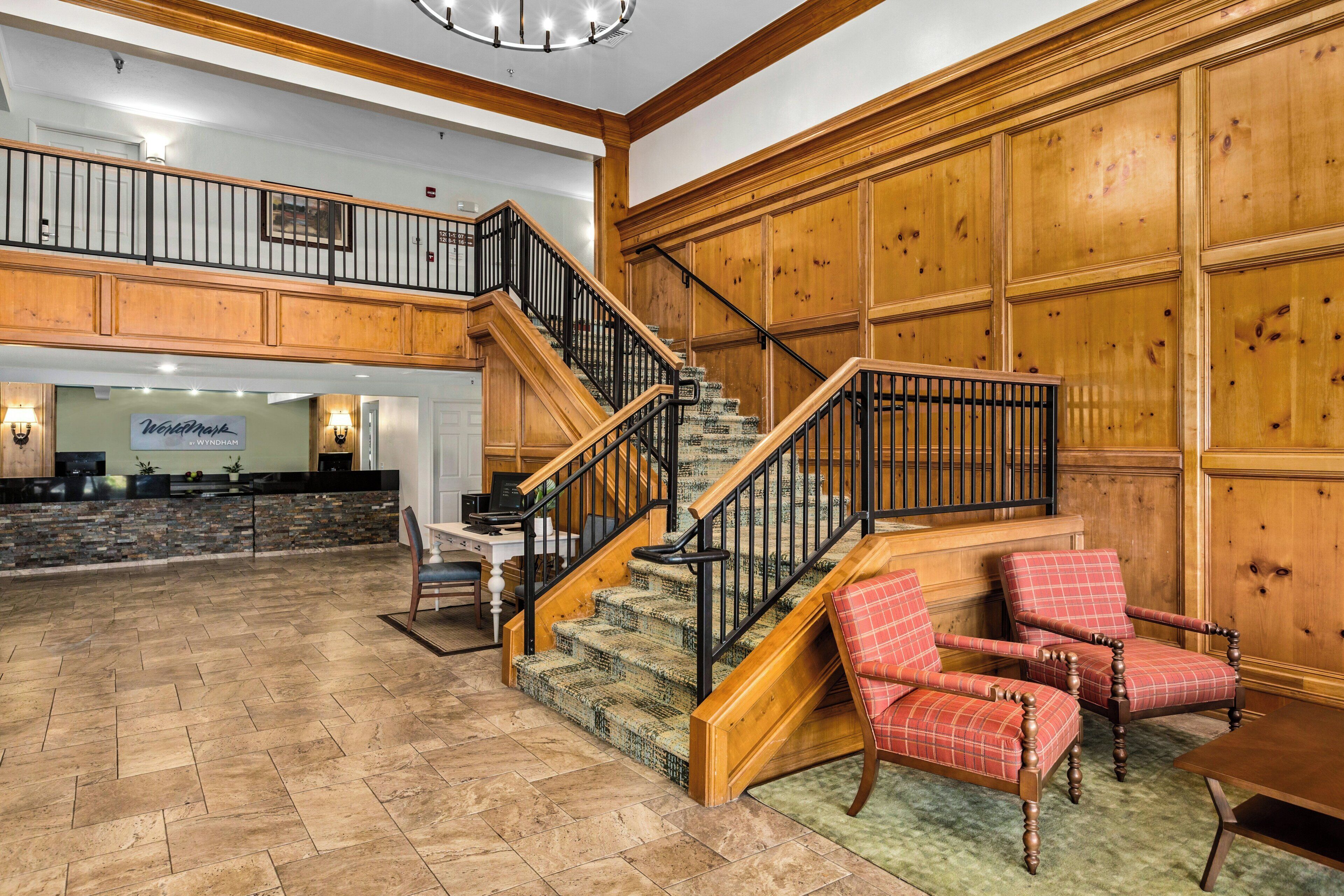 A spacious lobby with a wooden staircase, a reception desk, and a seating area with plaid chairs.