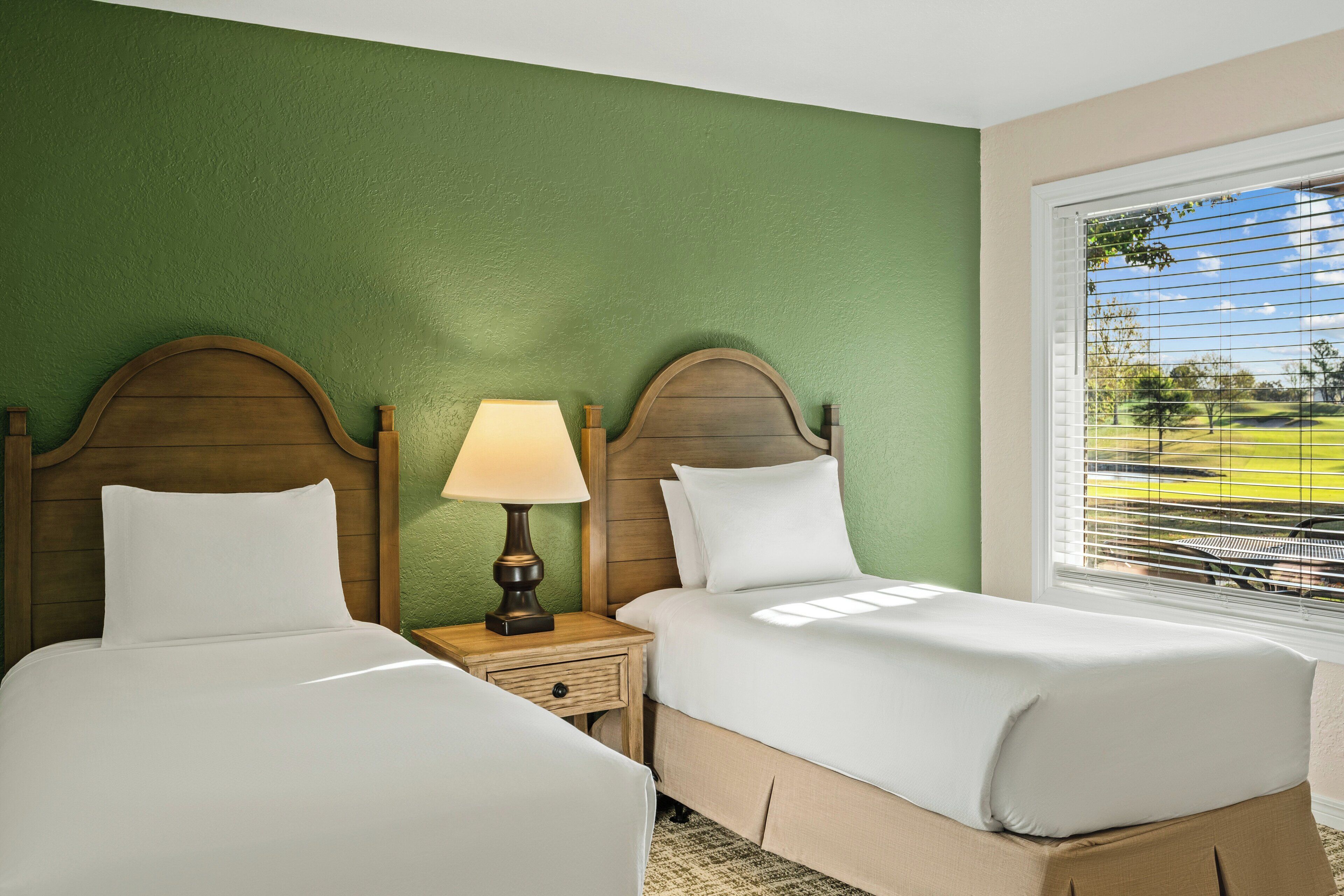 A hotel room with two beds, a nightstand with a lamp, and a window with blinds.
