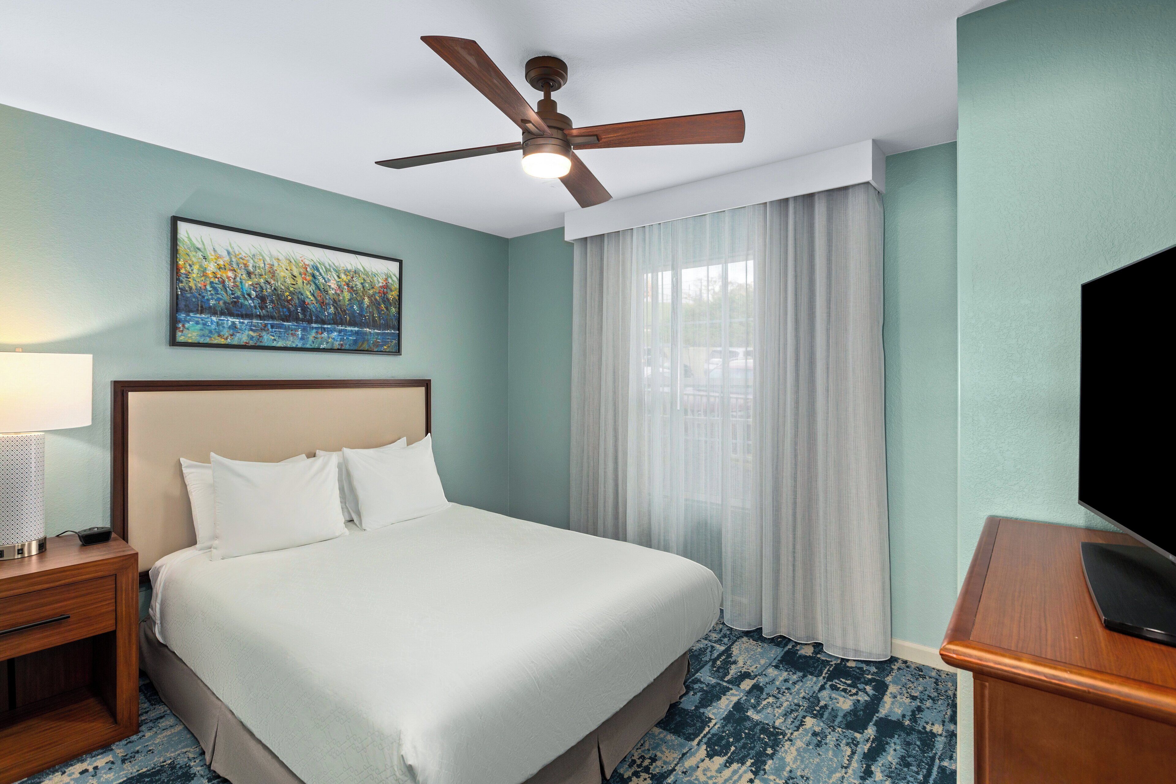 A hotel room with a bed, a ceiling fan, a painting, a TV, and a window with curtains.