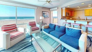 A modern living room with a large window overlooking the beach, a sofa, armchairs, a glass coffee table, and a kitchen area with wooden cabinets and a bar. - Resort at Seaside (Seaside)