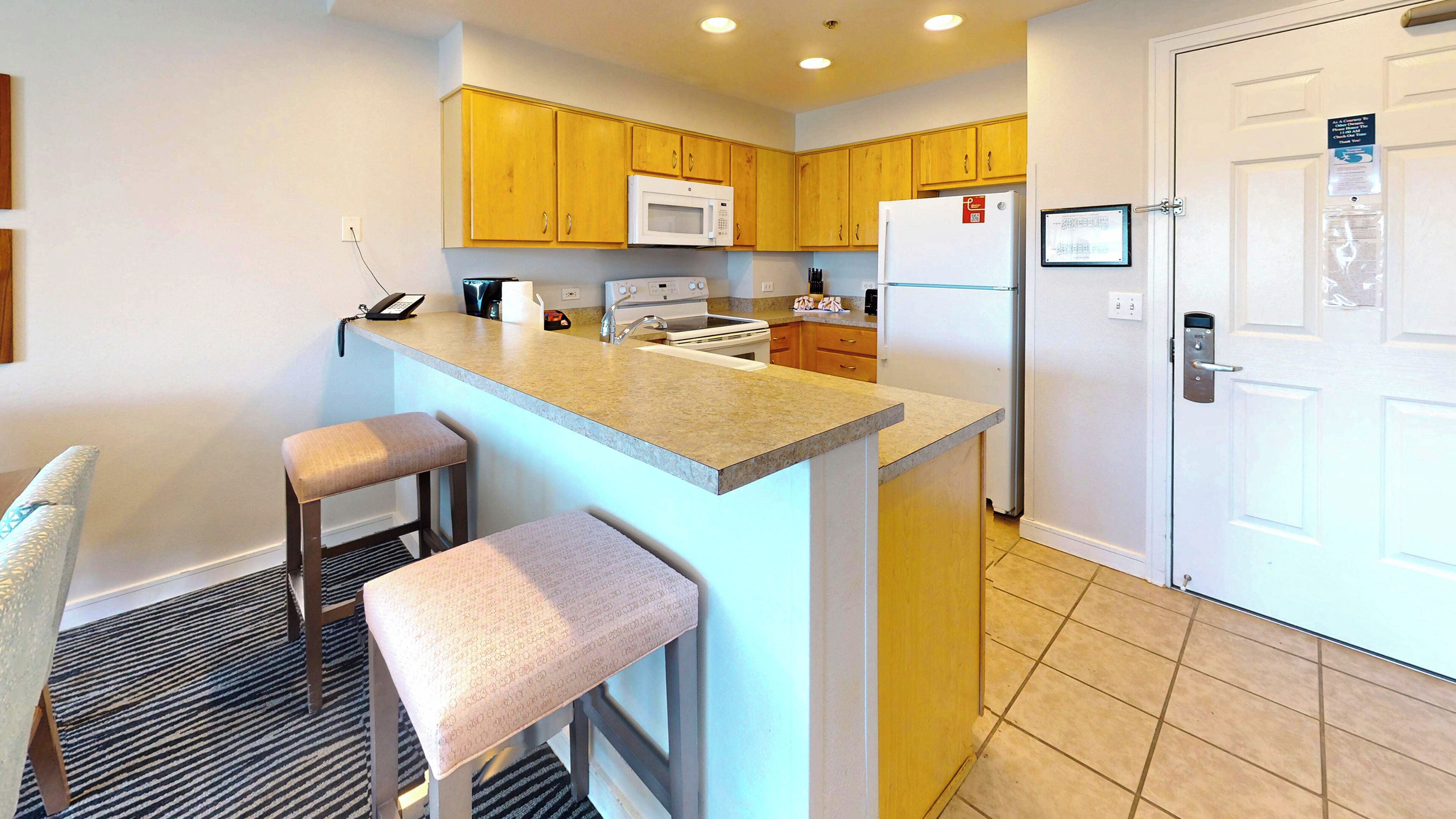 A modern kitchen with wooden cabinets, a white refrigerator, a microwave, and a built-in oven. There are two bar stools and a sink.