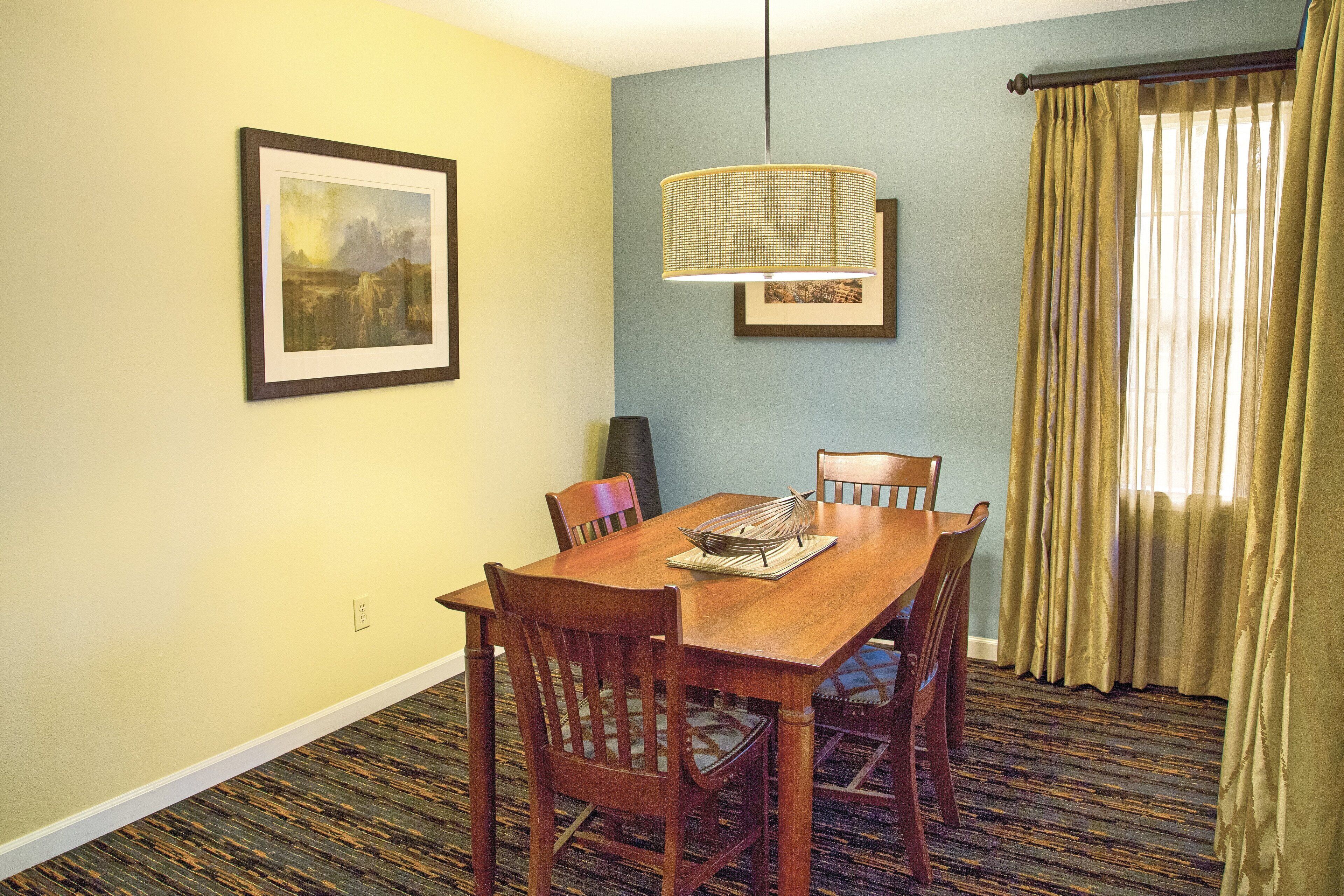 A dining area with a wooden table and chairs, a hanging lamp, and a framed picture on the wall.