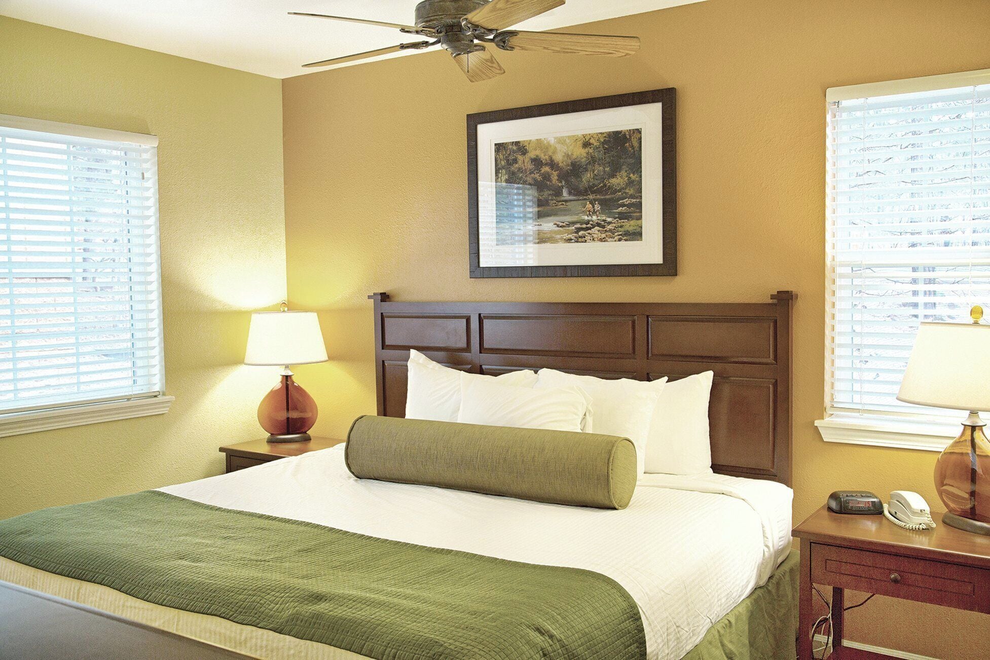 Une chambre d’hôtel avec un lit, un ventilateur de plafond, un tableau encadré au mur et un bureau avec une lampe et un téléphone.