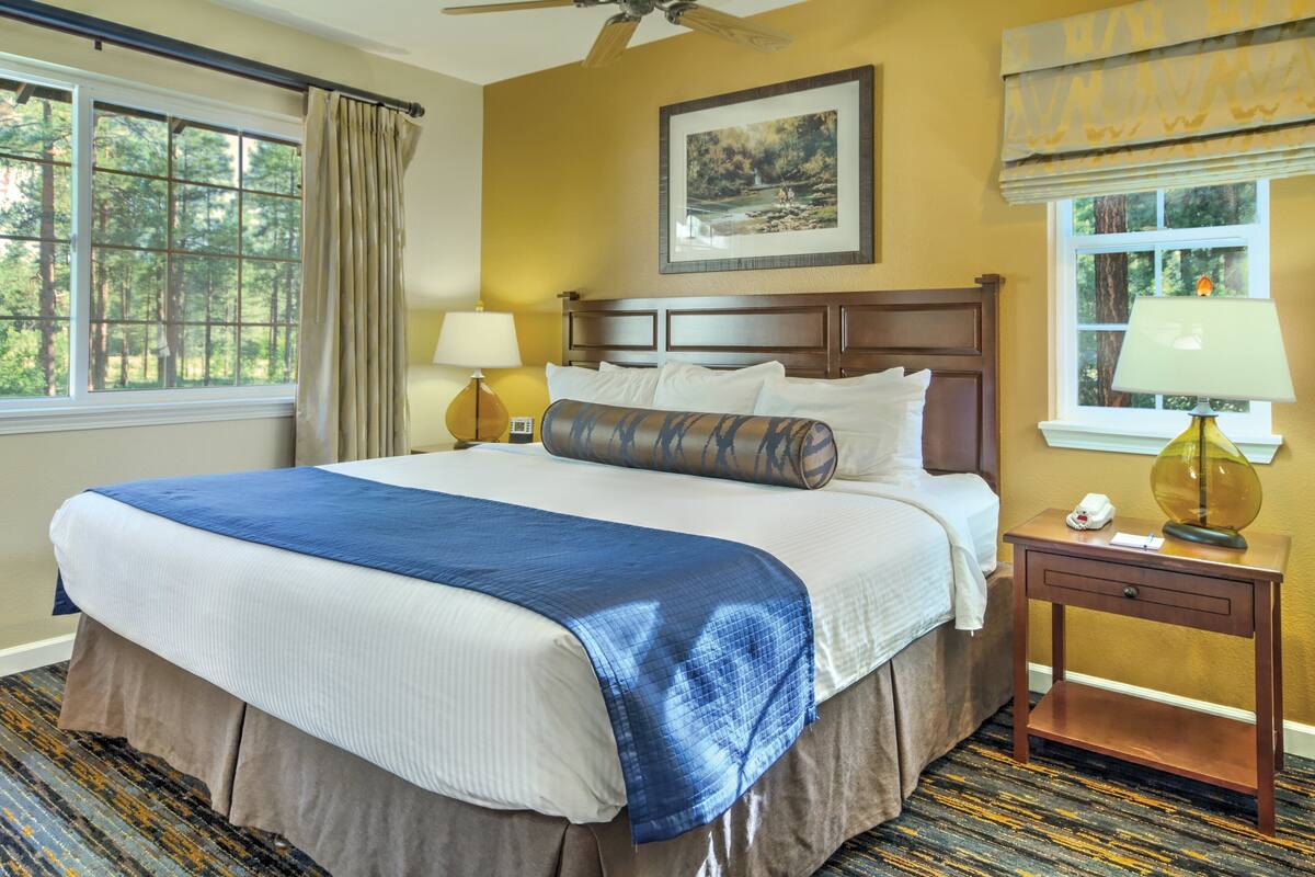 A hotel room with a large bed, a nightstand with a lamp, a window with curtains, and a ceiling fan.