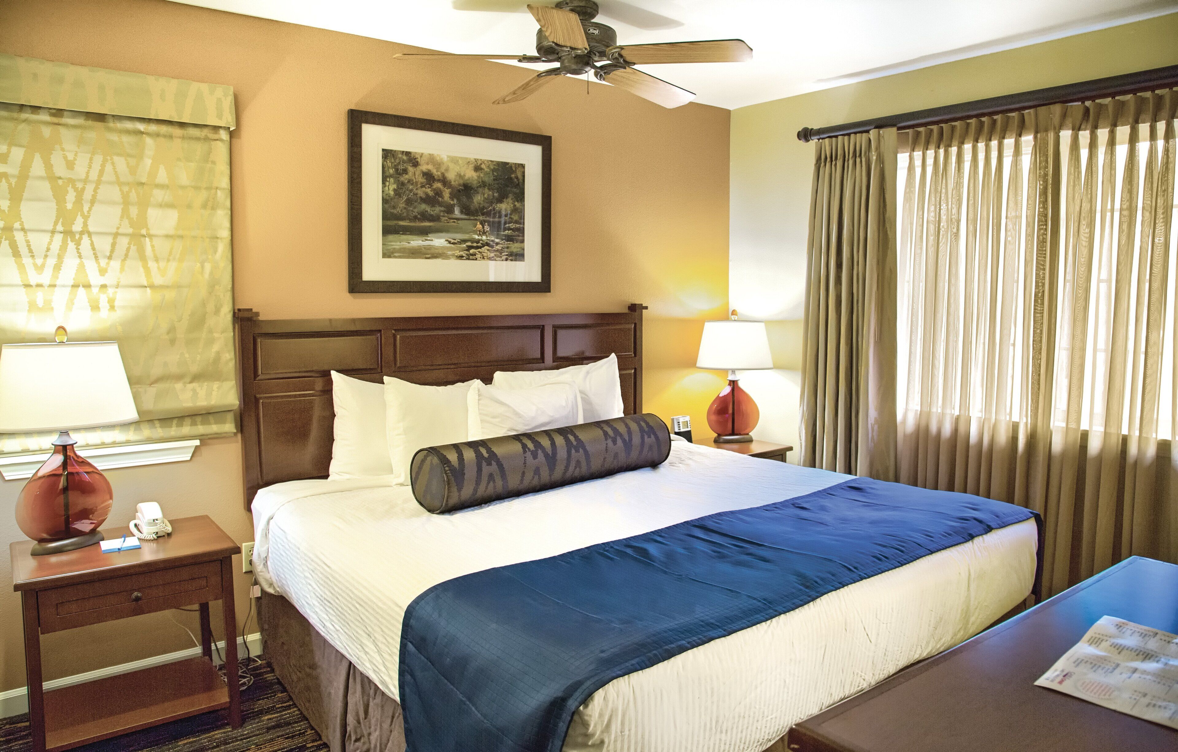 Quarto de hotel com cama, duas luminárias de cabeceira, um ventilador de teto, um quadro na parede e uma janela com cortinas.