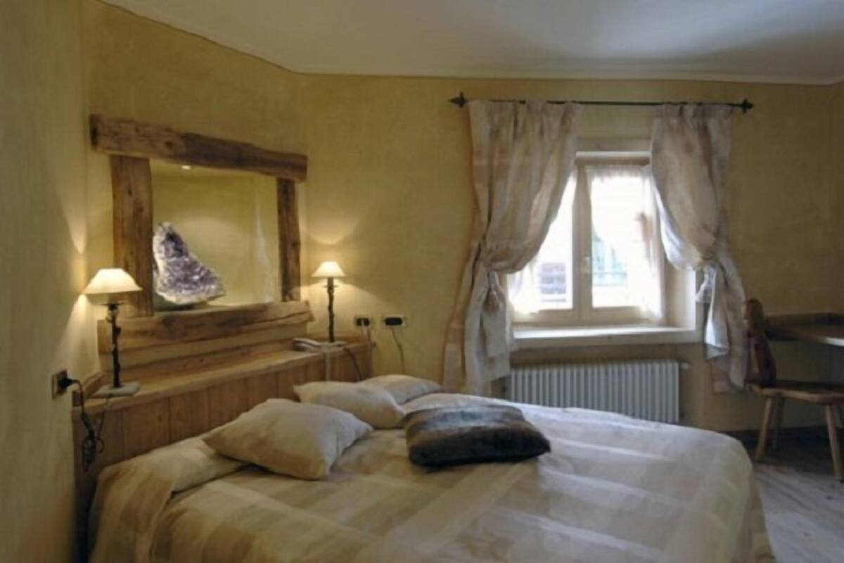 Ein Schlafzimmer mit Bett, Nachttisch, Lampe, Spiegel, Fenster mit Vorhängen und einem Sessel.