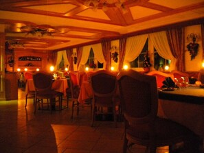Un restaurant à la lumière tamisée, avec des poutres en bois au plafond, des tables dressées pour le repas et des rideaux aux fenêtres.