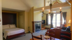 A hotel room with a bed, a fireplace, a dining table with chairs, and a chandelier. - Place St-Bernard (Mont-Tremblant)