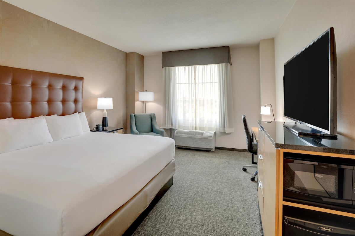 Ein Hotelzimmer mit einem großen Bett, einem Schreibtisch mit Flachbildfernseher, einem Sessel, einer Lampe und einem Fenster mit Vorhängen.