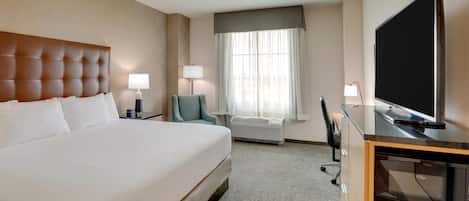 A hotel room with a large bed, a desk with a flat-screen TV, a chair, a lamp, and a window with curtains.
