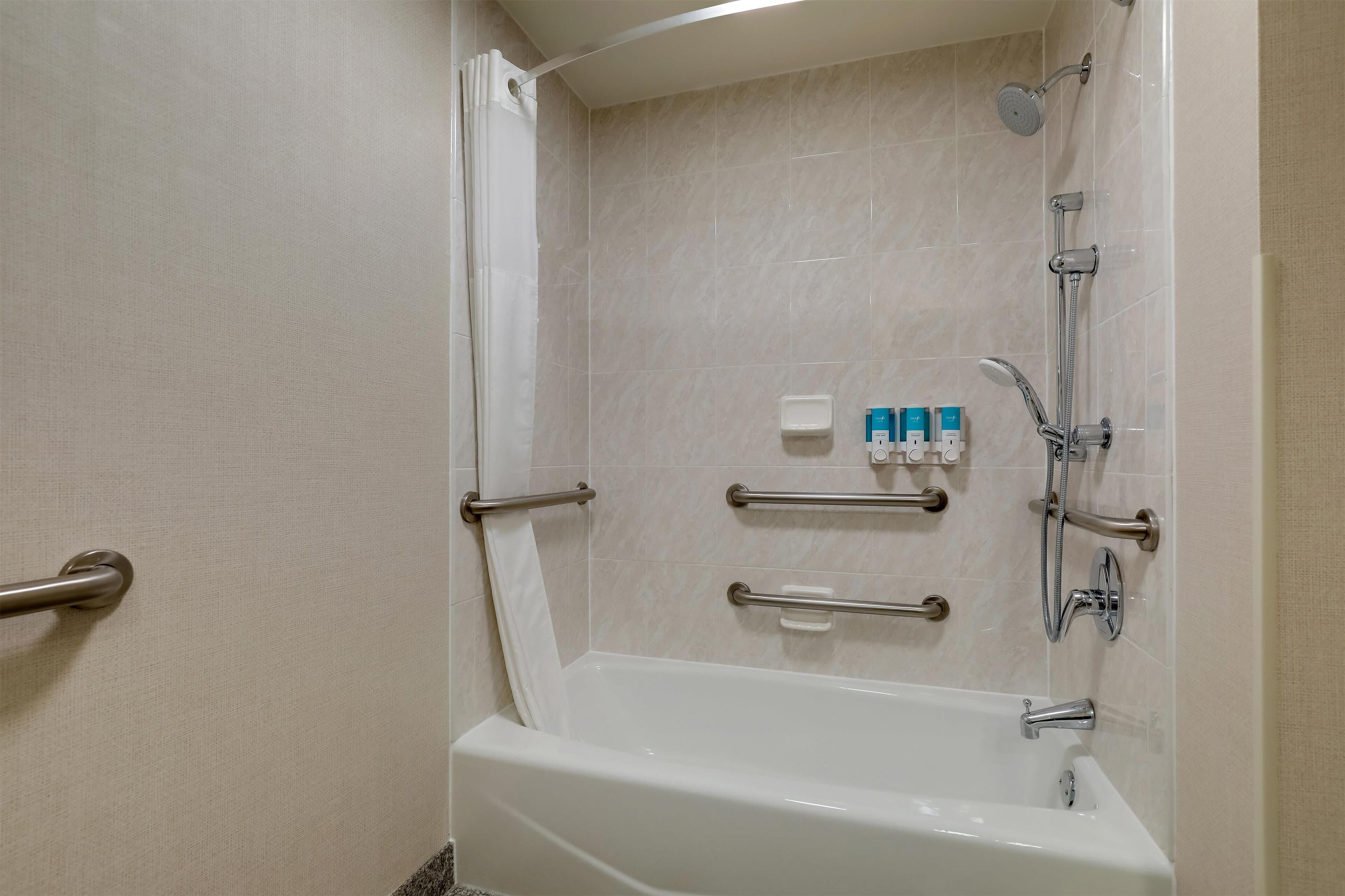 A bathroom with a bathtub, grab bars, a showerhead, and a soap dispenser.